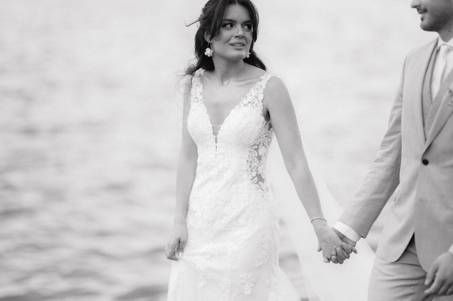 A couple in wedding attire holds hands by the water in an elegant black and white photograph.