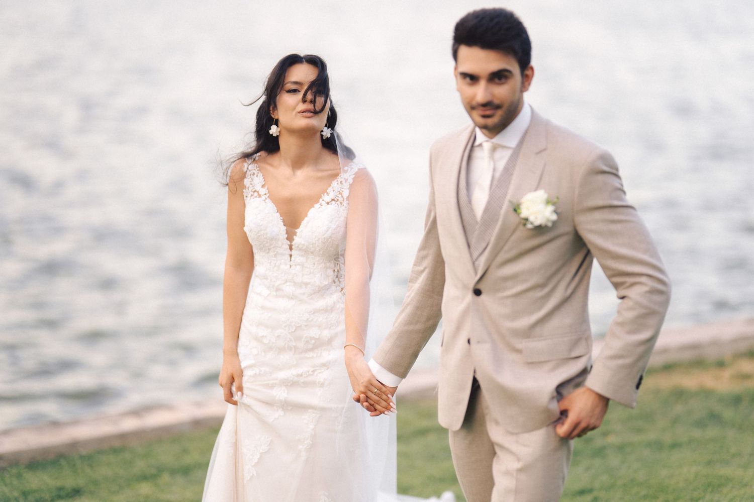 A couple in wedding attire holds hands while walking by the water, with the bride in lace and groom in beige suit.