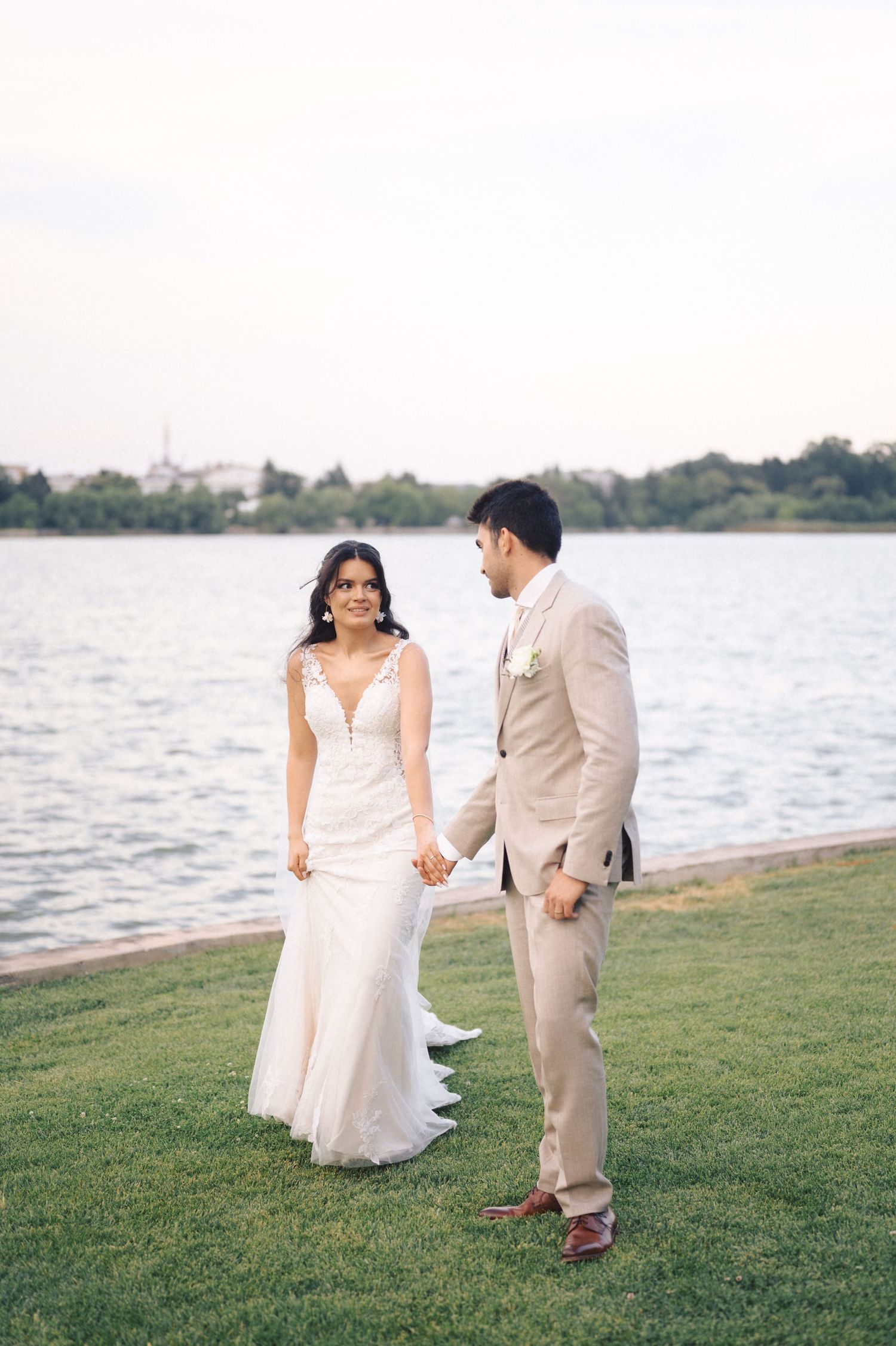 A romantic wedding photoshoot by the waterfront with a couple walking on grass in elegant attire.