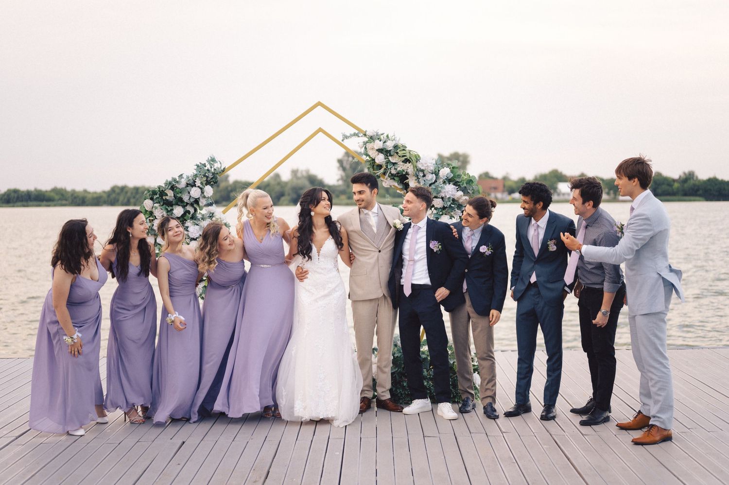 Wedding party in purple bridesmaid dresses and navy suits pose together on wooden dock by waterfront under geometric arch.