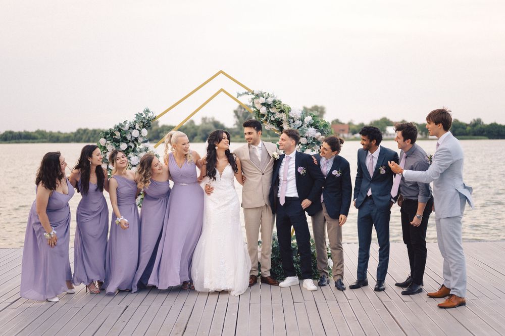 Wedding party in purple bridesmaid dresses and navy suits pose together on wooden dock by waterfront under geometric arch.