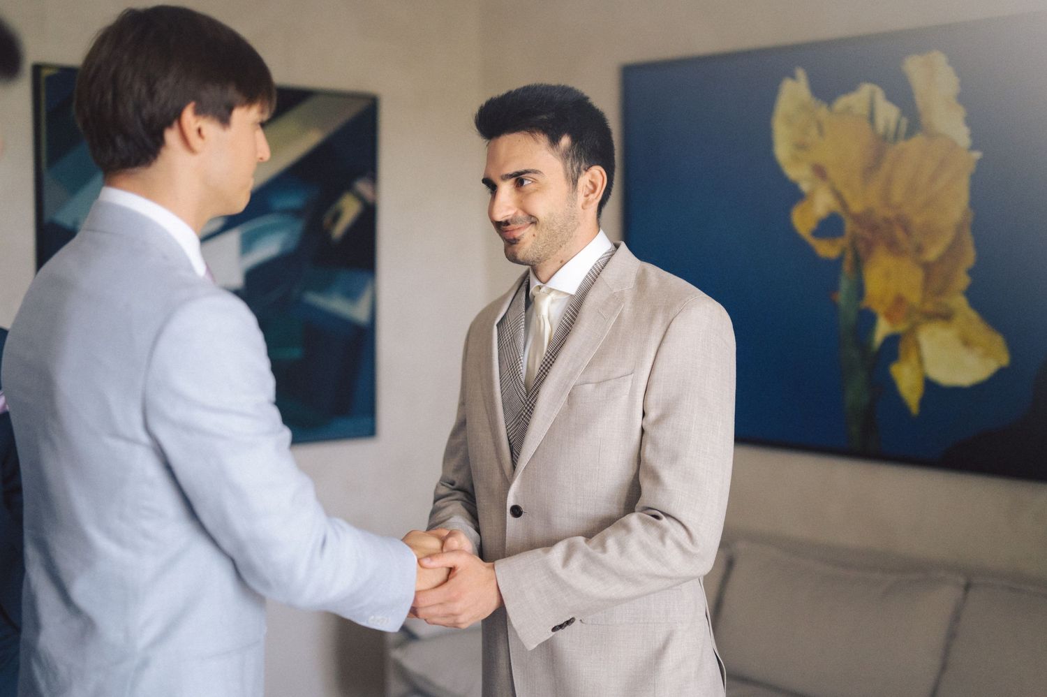 Two professionals in light colored suits shake hands in an office space with blue floral artwork on the walls.