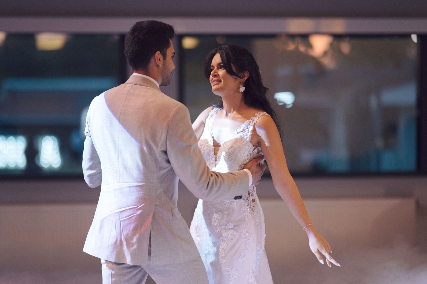 A couple dressed in elegant white attire dances together on a dimly lit dance floor during a special occasion.