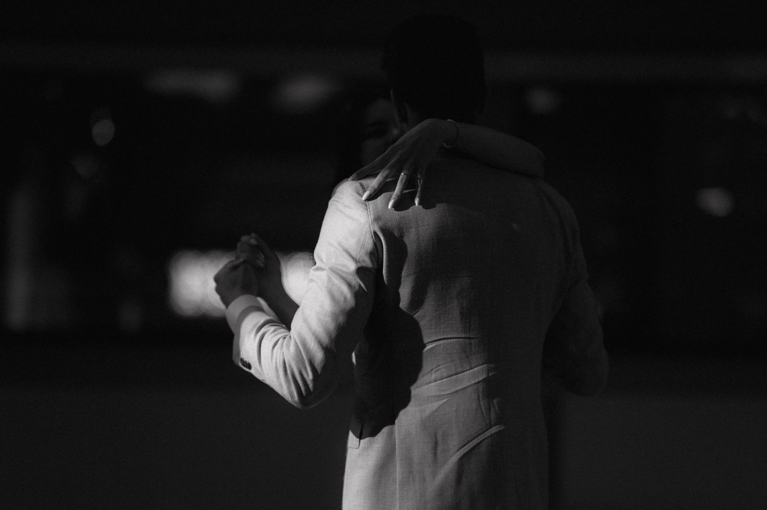 Black and white photo of a tender dance moment with two figures embracing in dramatic lighting on a dark stage.