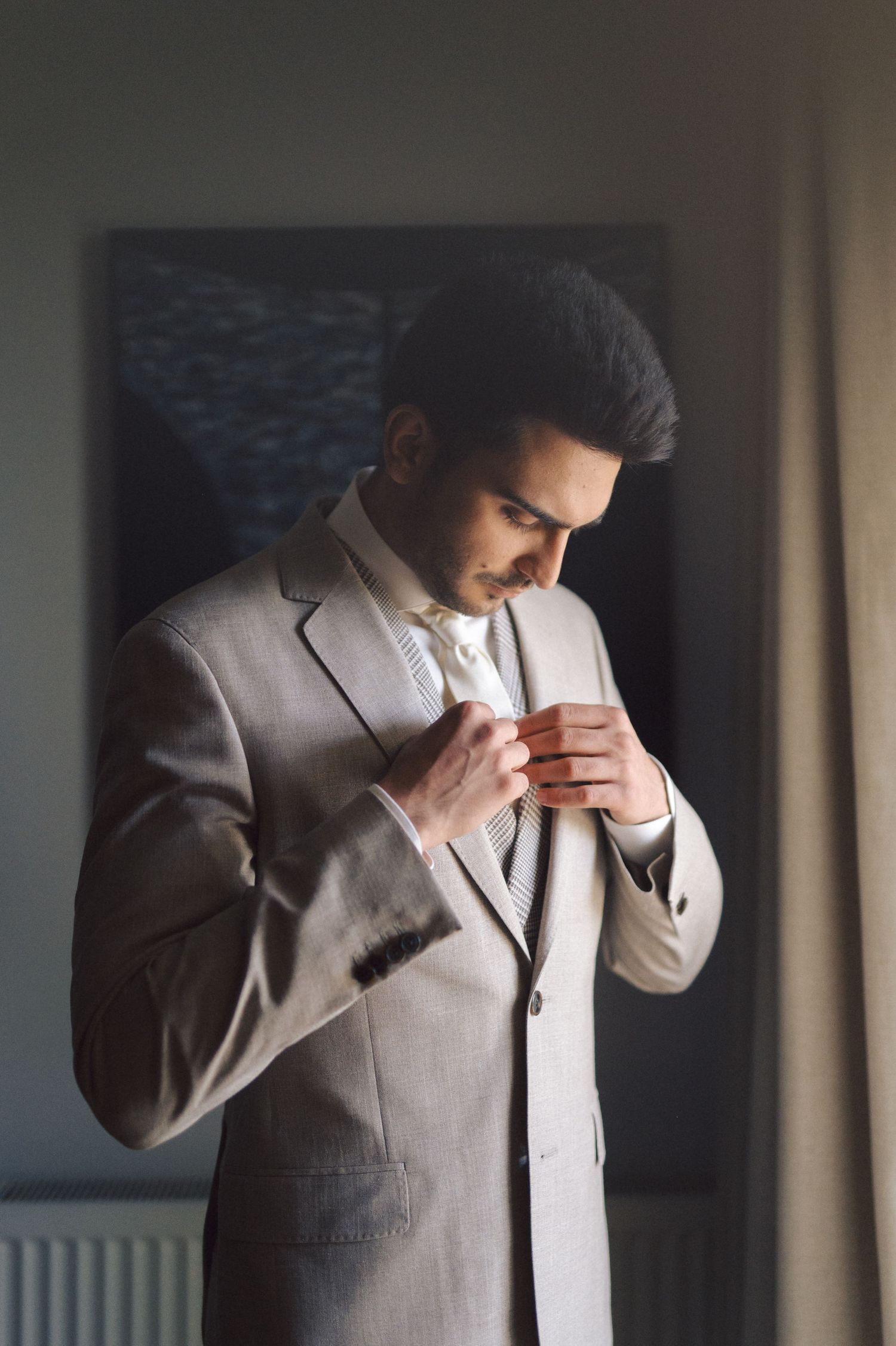 A person in a light gray suit adjusts their tie while standing in a dimly lit room near a window.