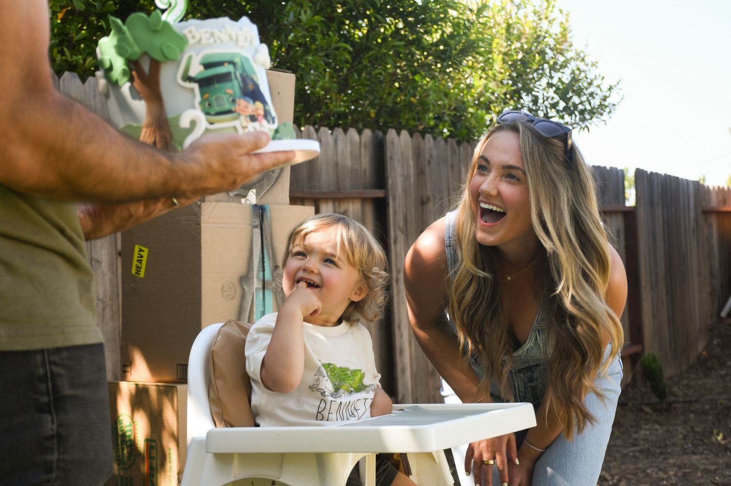 Mother and toddler laughing joyfully at a birthday cake in a sunny backyard setting.