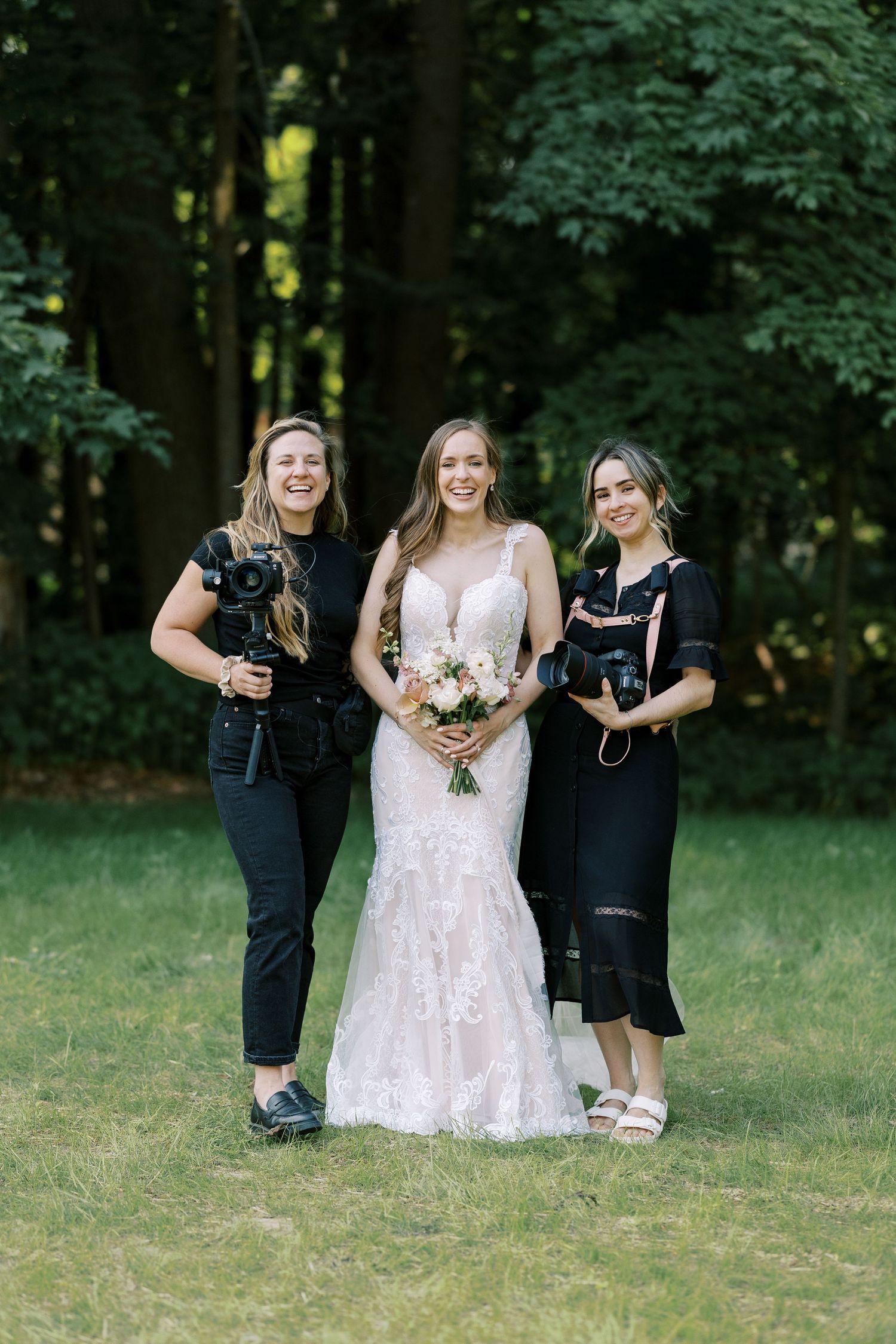 Professional photographers in black outfits with cameras stand alongside a bride holding a pastel bouquet in outdoor setting.