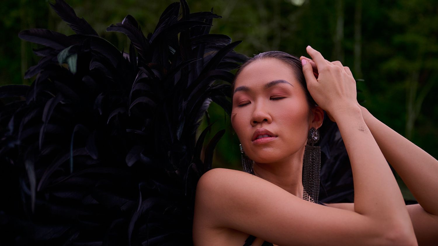 A figure in a dark feathered outfit poses dramatically against a natural green backdrop in soft lighting.