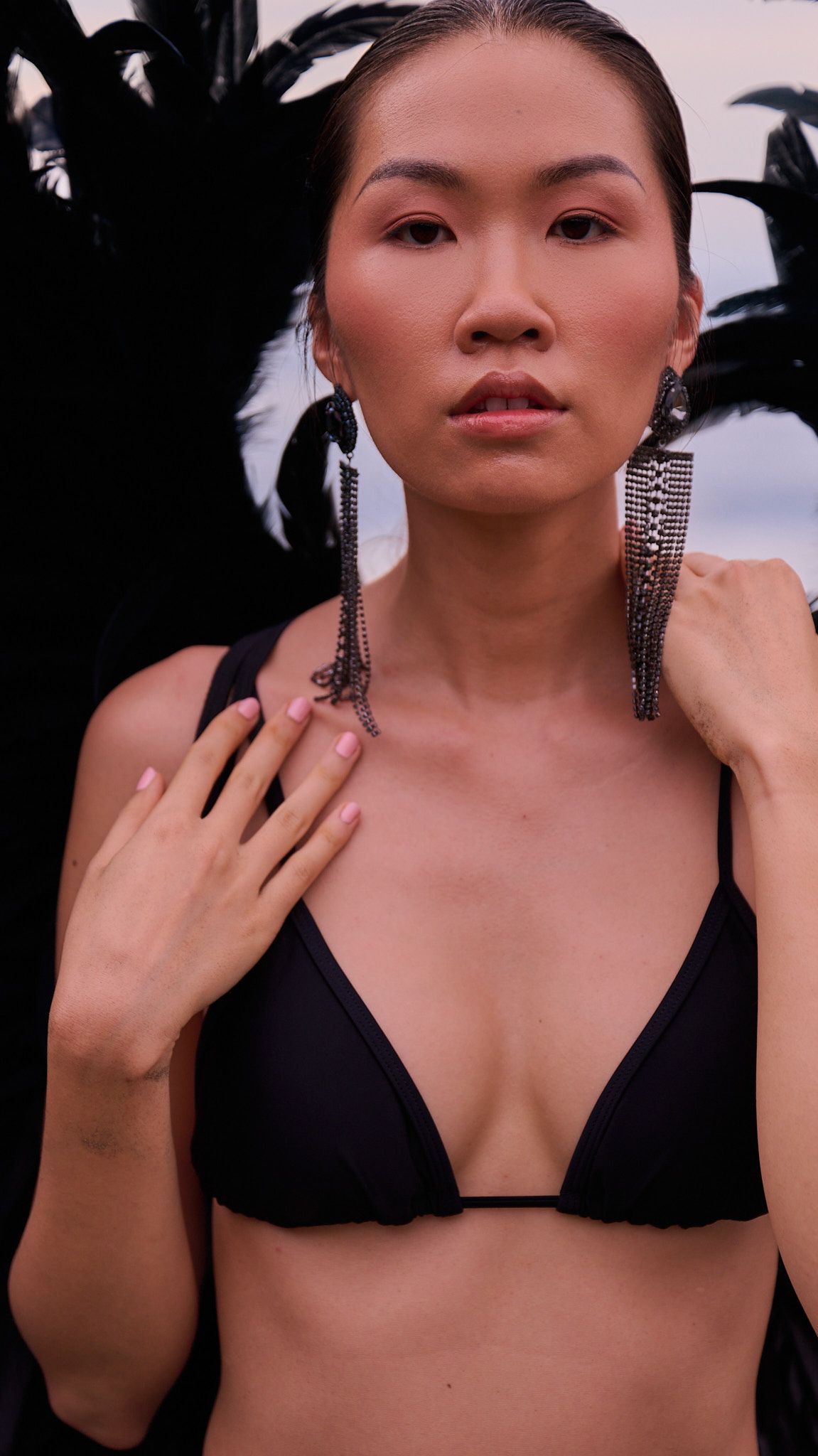 Model wearing a black bikini top and dangling silver statement earrings poses against dark foliage background.