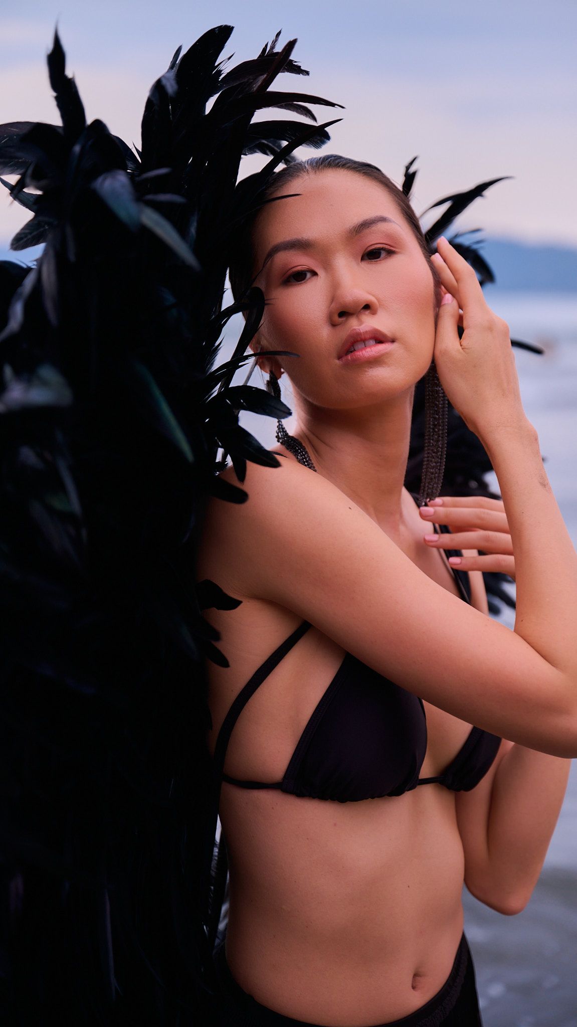Fashion portrait featuring black feather accessories and swimwear against purple sunset sky and ocean.
