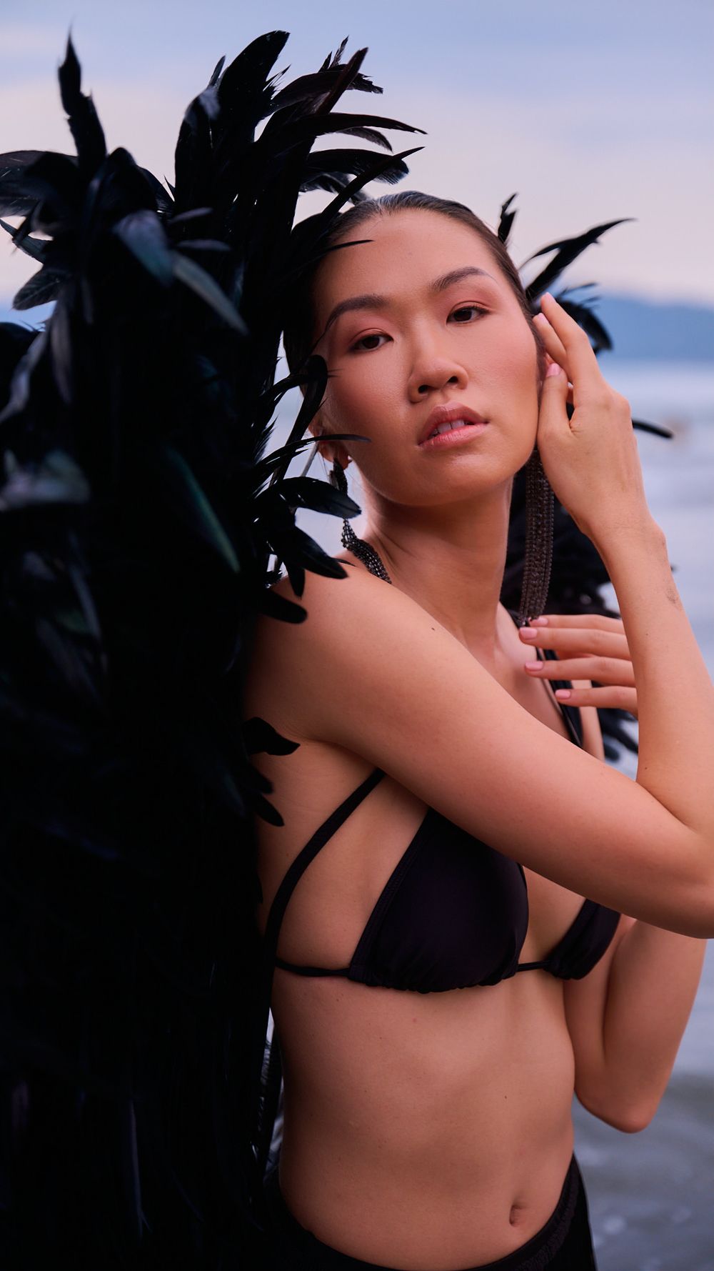 Fashion portrait featuring black feather accessories and swimwear against purple sunset sky and ocean.