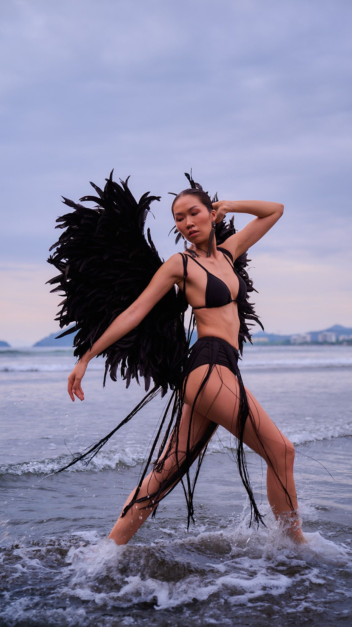 A dancer in black swimwear and feathers strikes dynamic poses in the ocean waves against a cloudy sky.