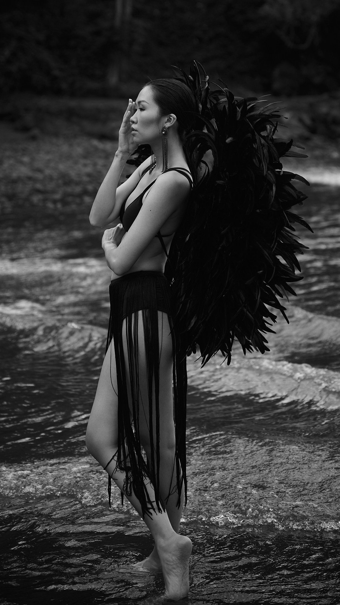 A figure in a dark dress with dramatic feathers stands in flowing water creating a moody black and white artistic scene.