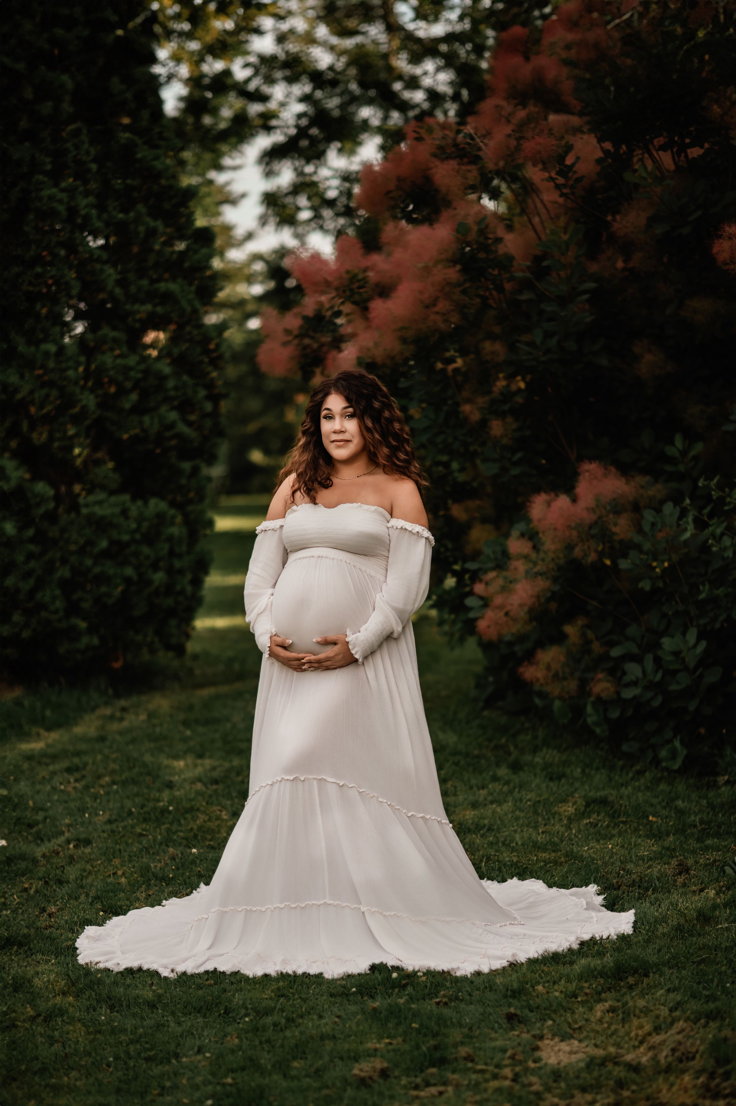 A maternity photoshoot in a park setting showing a white off-shoulder dress against autumn foliage in the background.
