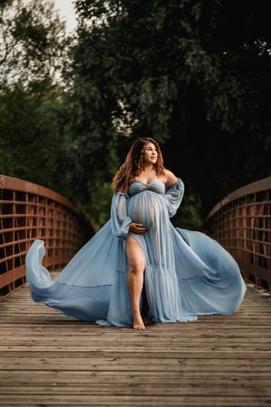 Flowing light blue maternity gown billows dramatically on wooden bridge surrounded by green trees.
