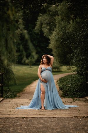 Expectant mother in flowing light blue gown poses gracefully on wooden bridge surrounded by lush summer greenery.