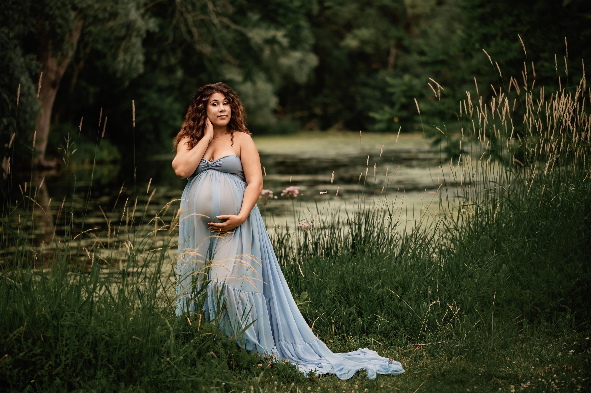 A woman in a flowing light blue dress poses gracefully in a lush outdoor garden setting during a photoshoot at sunset.