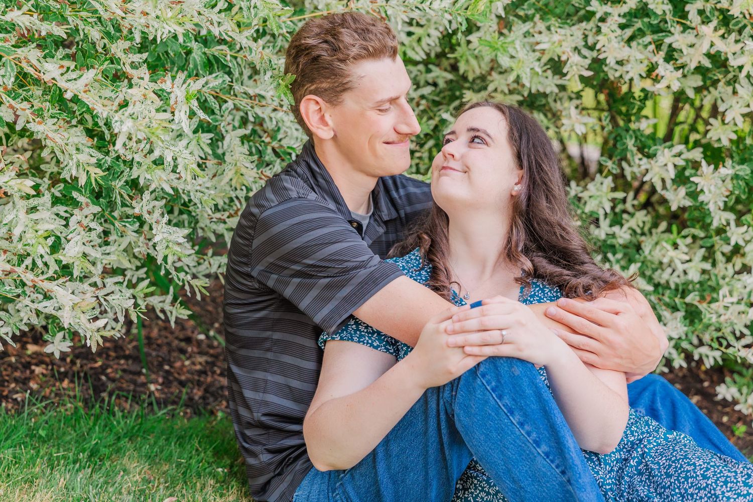 A romantic couple embraces while sitting on grass against a backdrop of flowering bushes in springtime.