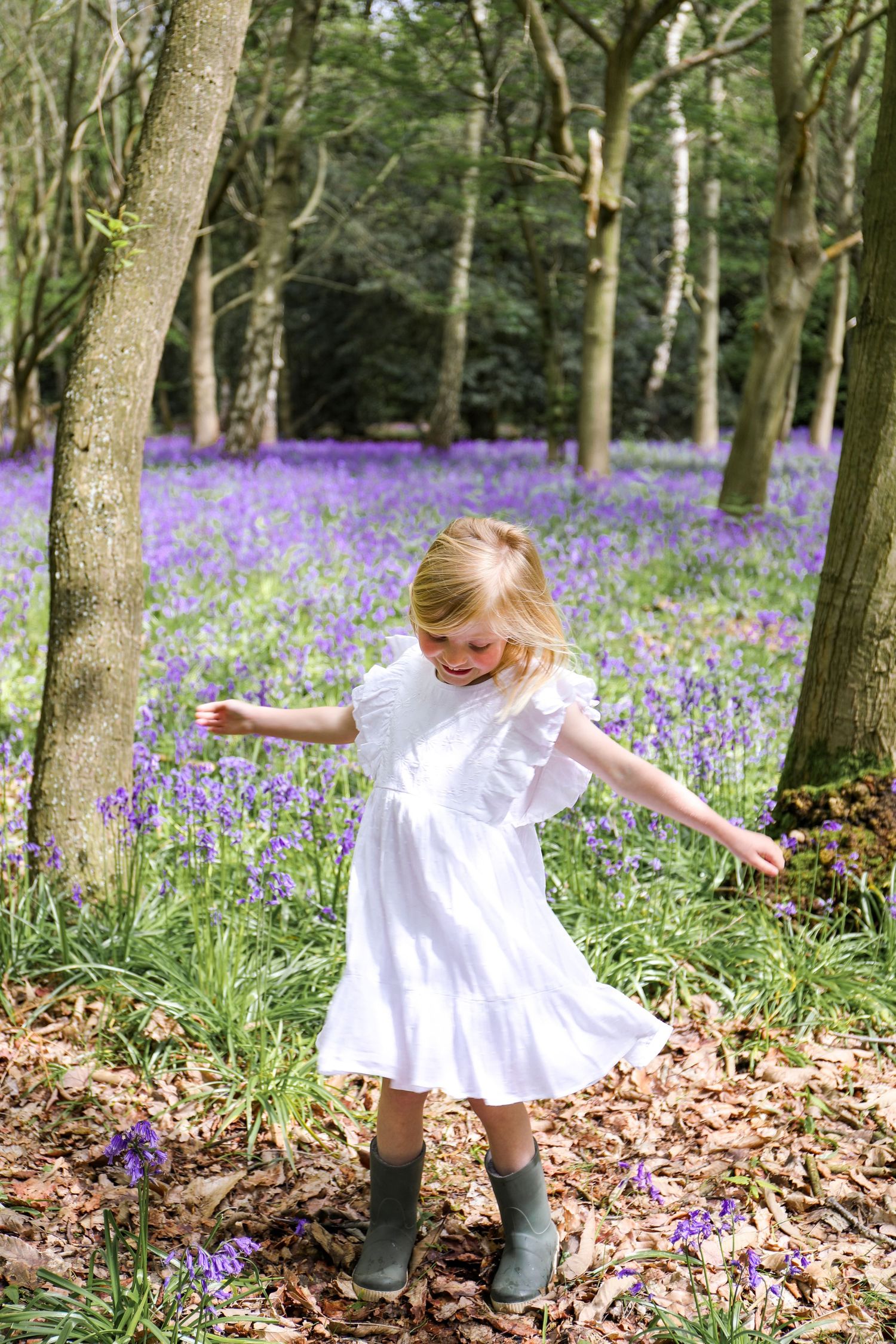 Girl in white dress twirls joyfully among vibrant purple bluebells in a spring woodland setting, during family shoot in Nottingham