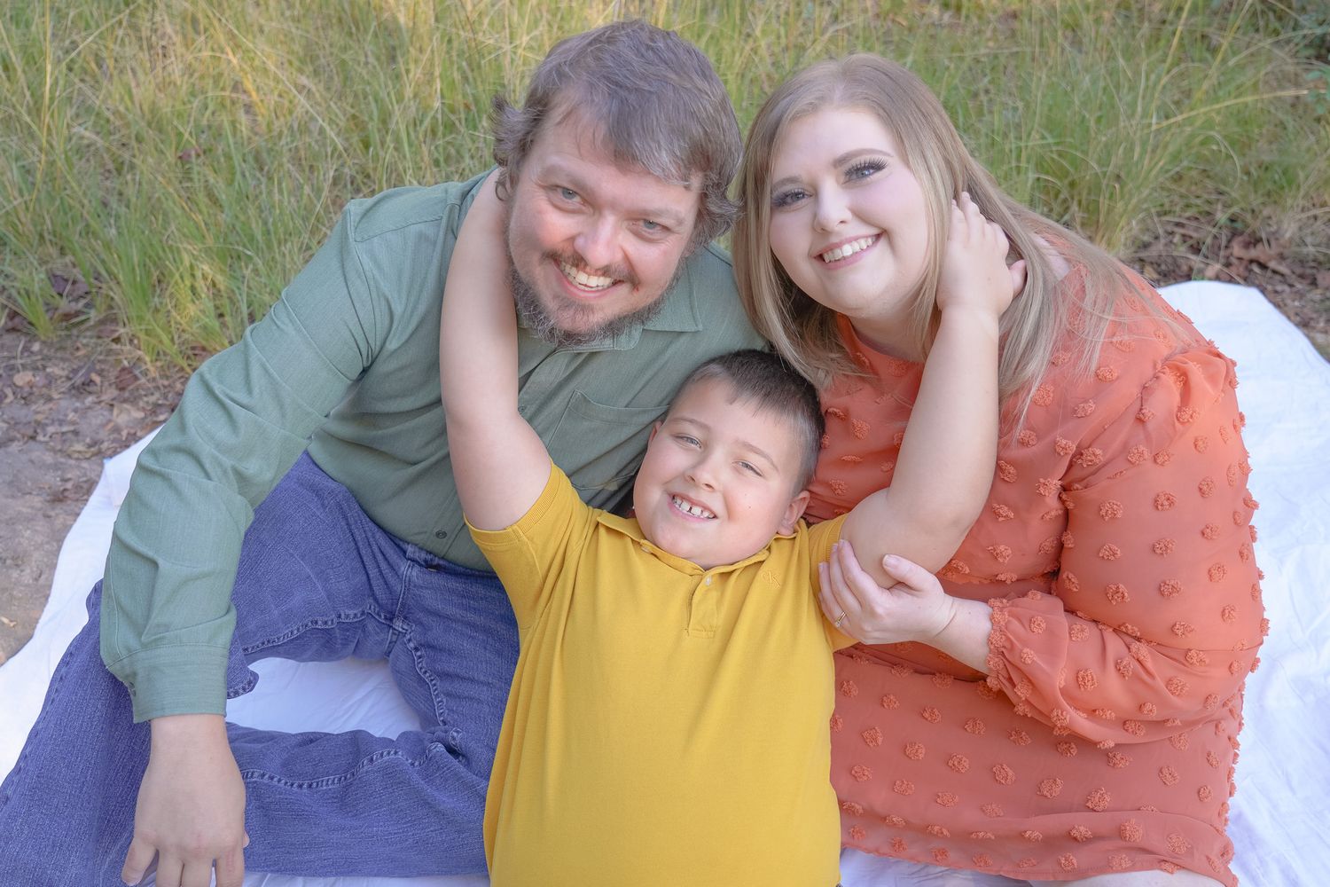 A happy family of three wearing green, yellow, and orange sits together on a blanket outdoors. Lincolnton NC Family Photography