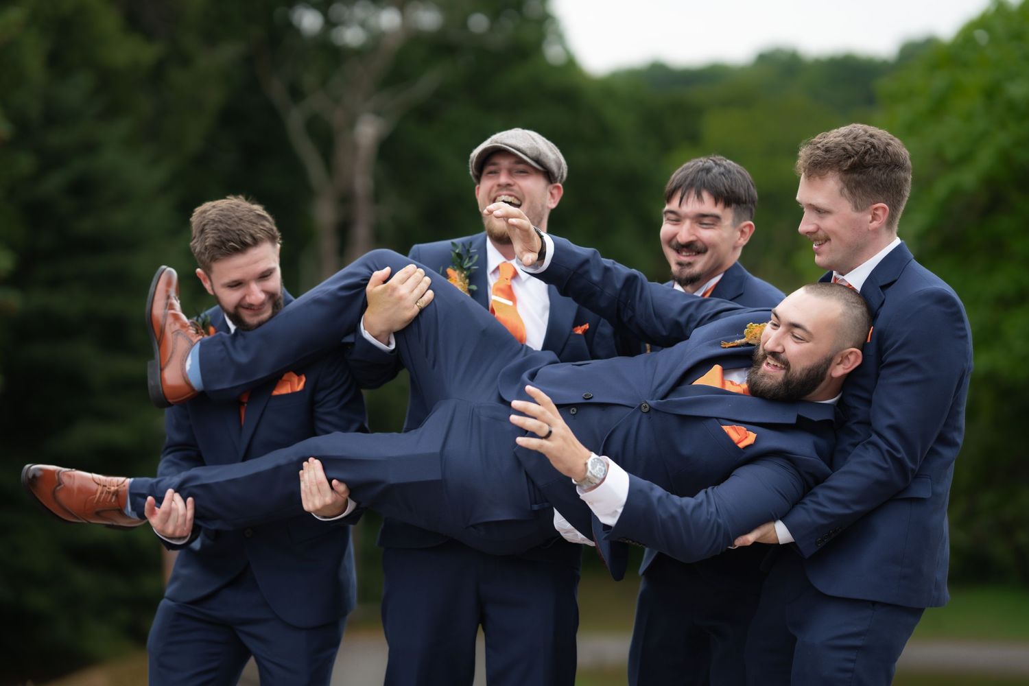 Groomsmen in navy suits playfully lift someone while celebrating outdoors.