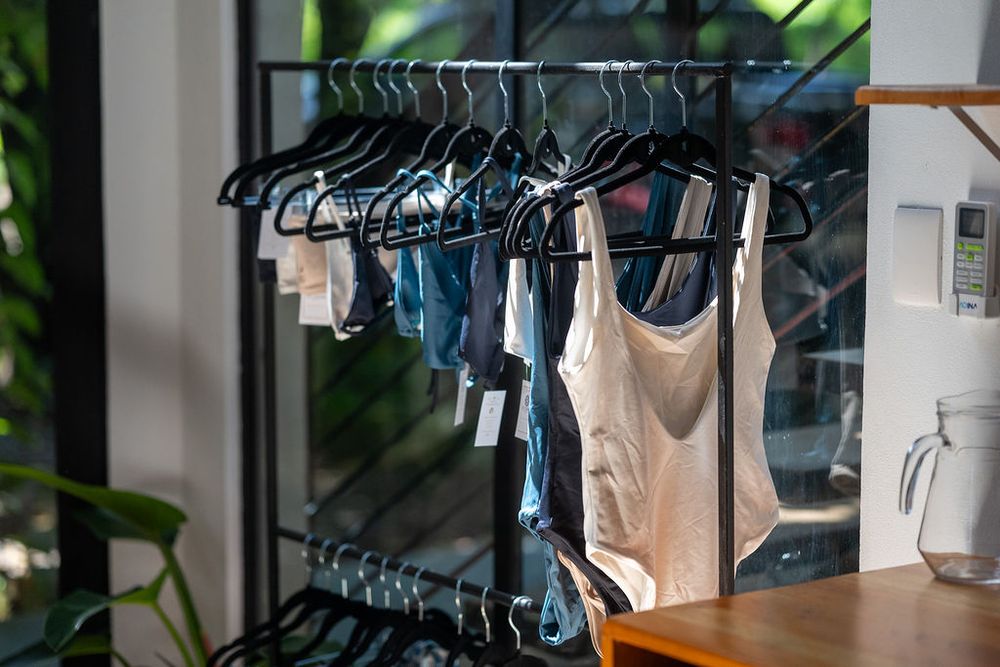 Clothing rack displaying denim jeans and tank tops with black hangers against a white wall with greenery.