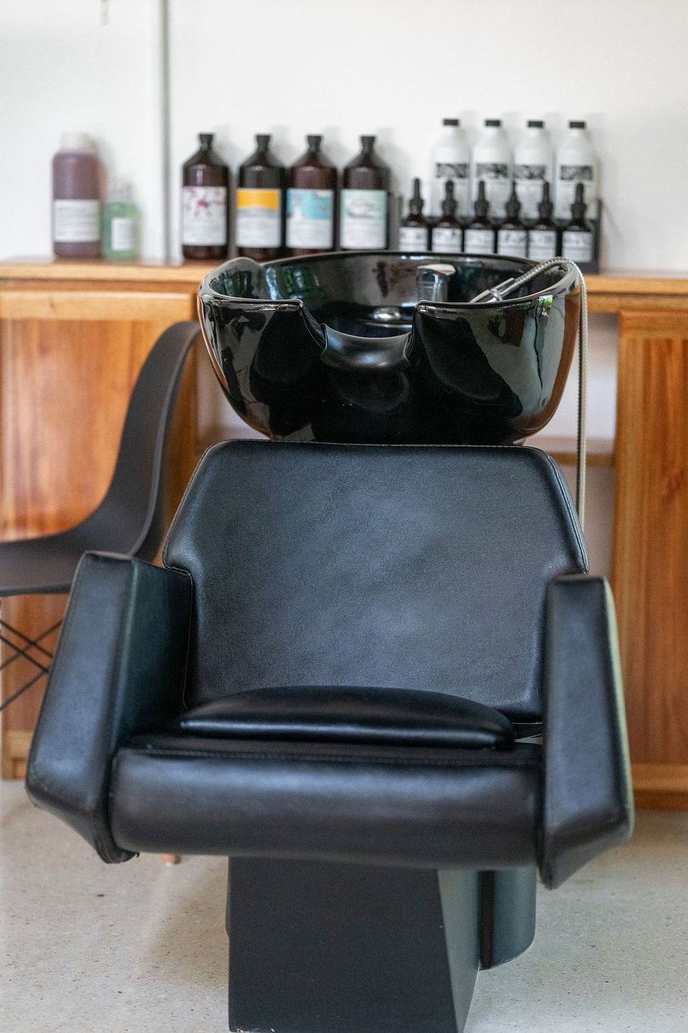 Professional salon hair washing station with black leather chair and ceramic sink with product bottles displayed on wooden shelf.