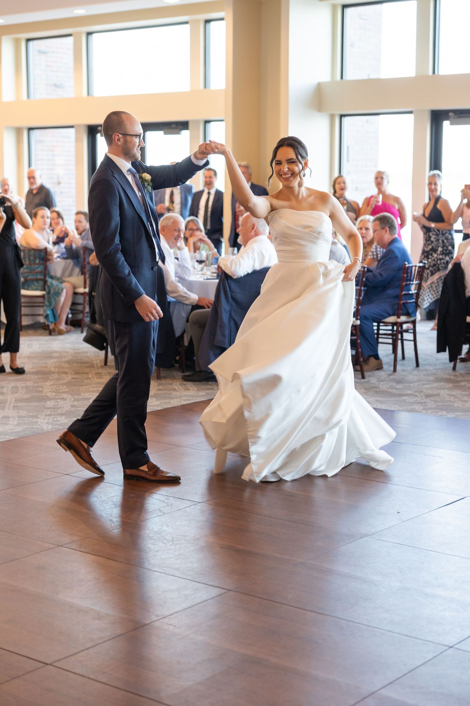Newlyweds share their first dance on a hardwood floor while wedding guests look on. Wedding Photographer Rachel Martinez Photography
