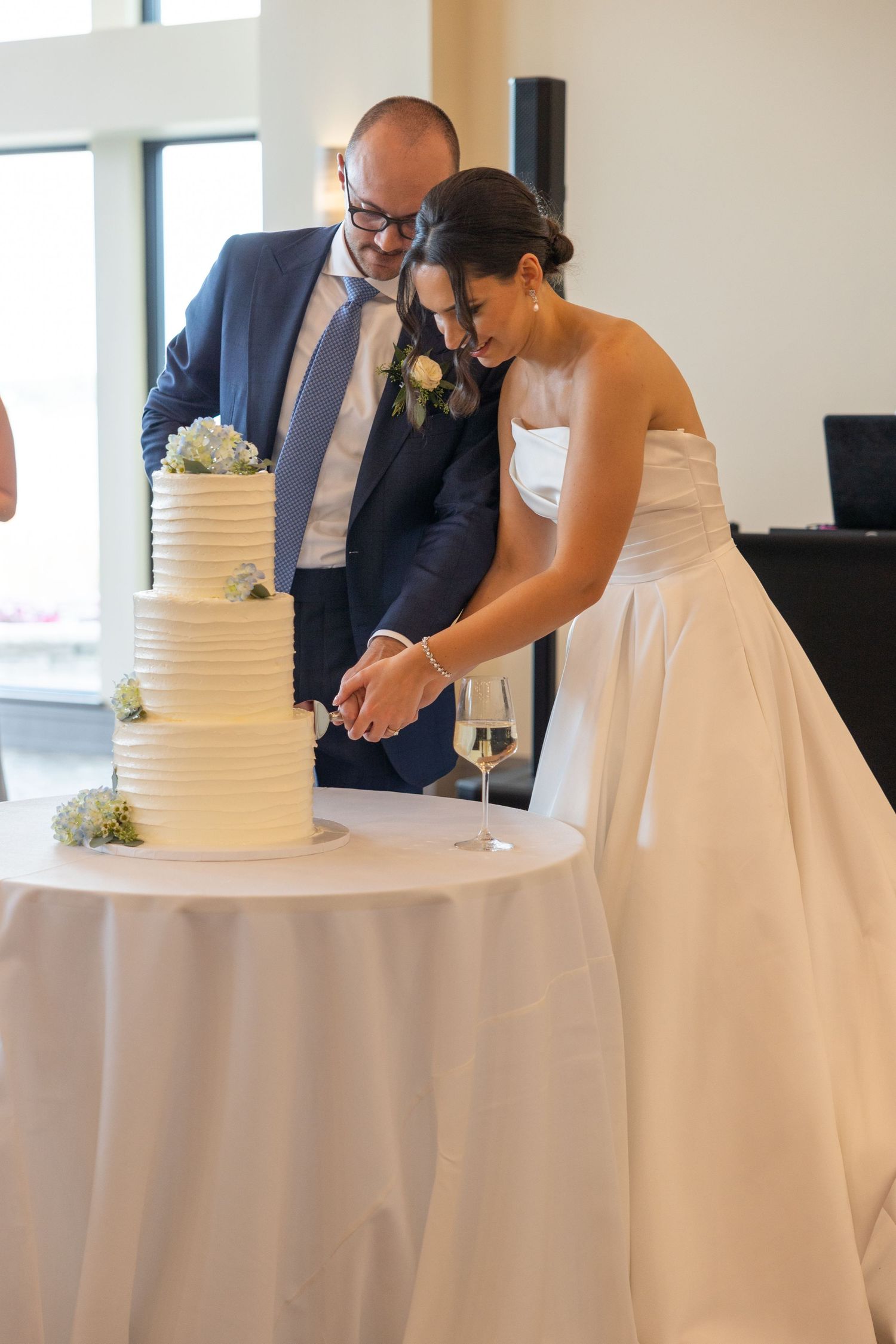 Newlyweds cut their elegant three-tiered white wedding cake decorated with flowers at a bright reception venue in Metro Detroit, Michigan