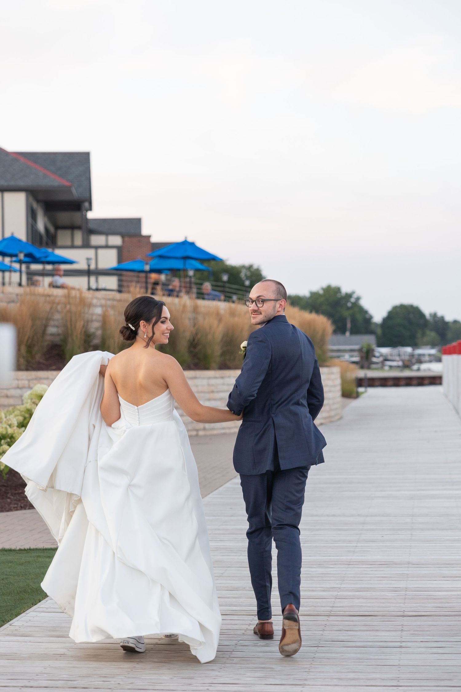 A couple in white wedding dress and navy suit walks away along a waterfront boardwalk during their wedding in Metro Detroit.