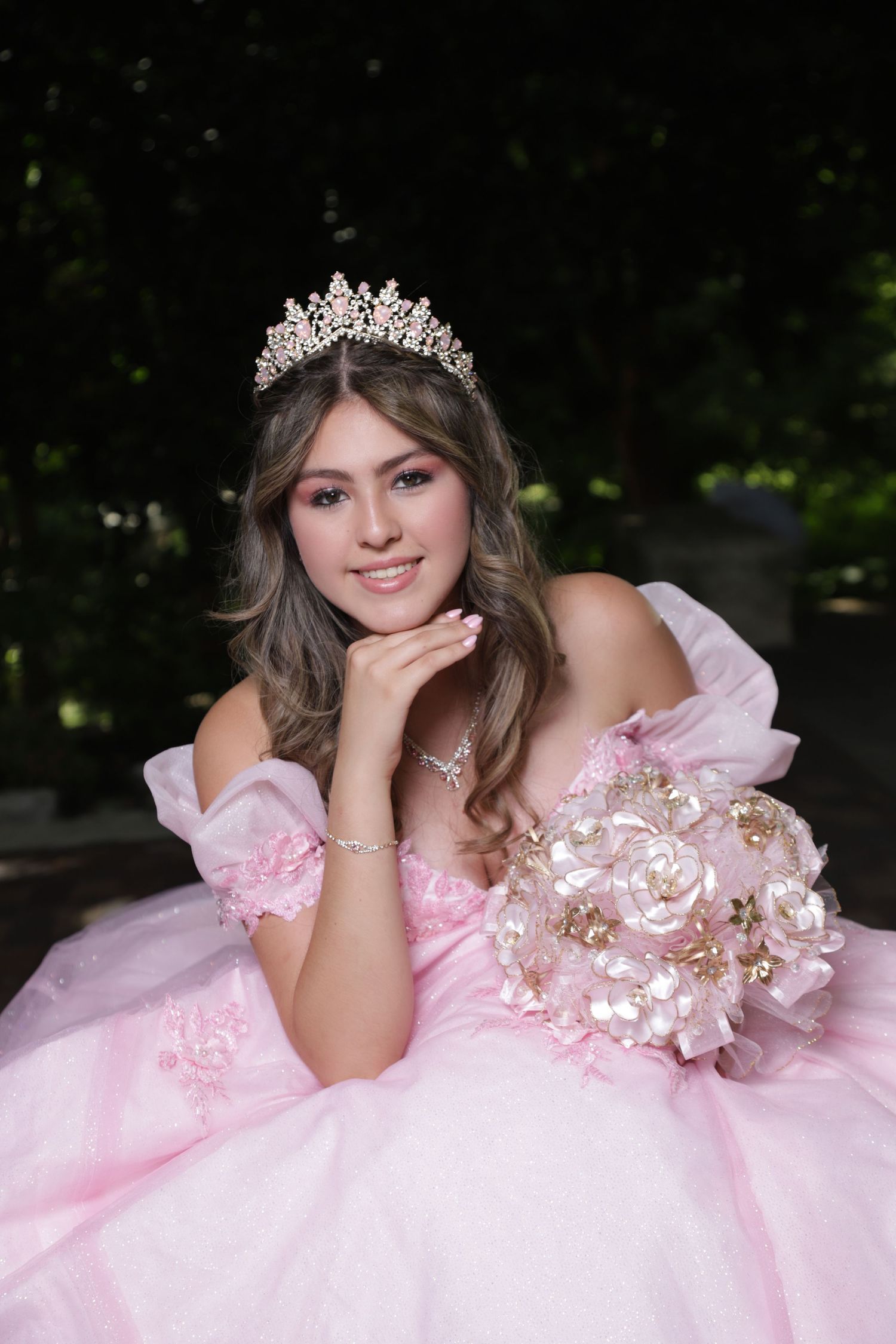 A person in a pink off-shoulder ball gown and crystal tiara poses with a floral bouquet in an outdoor setting.