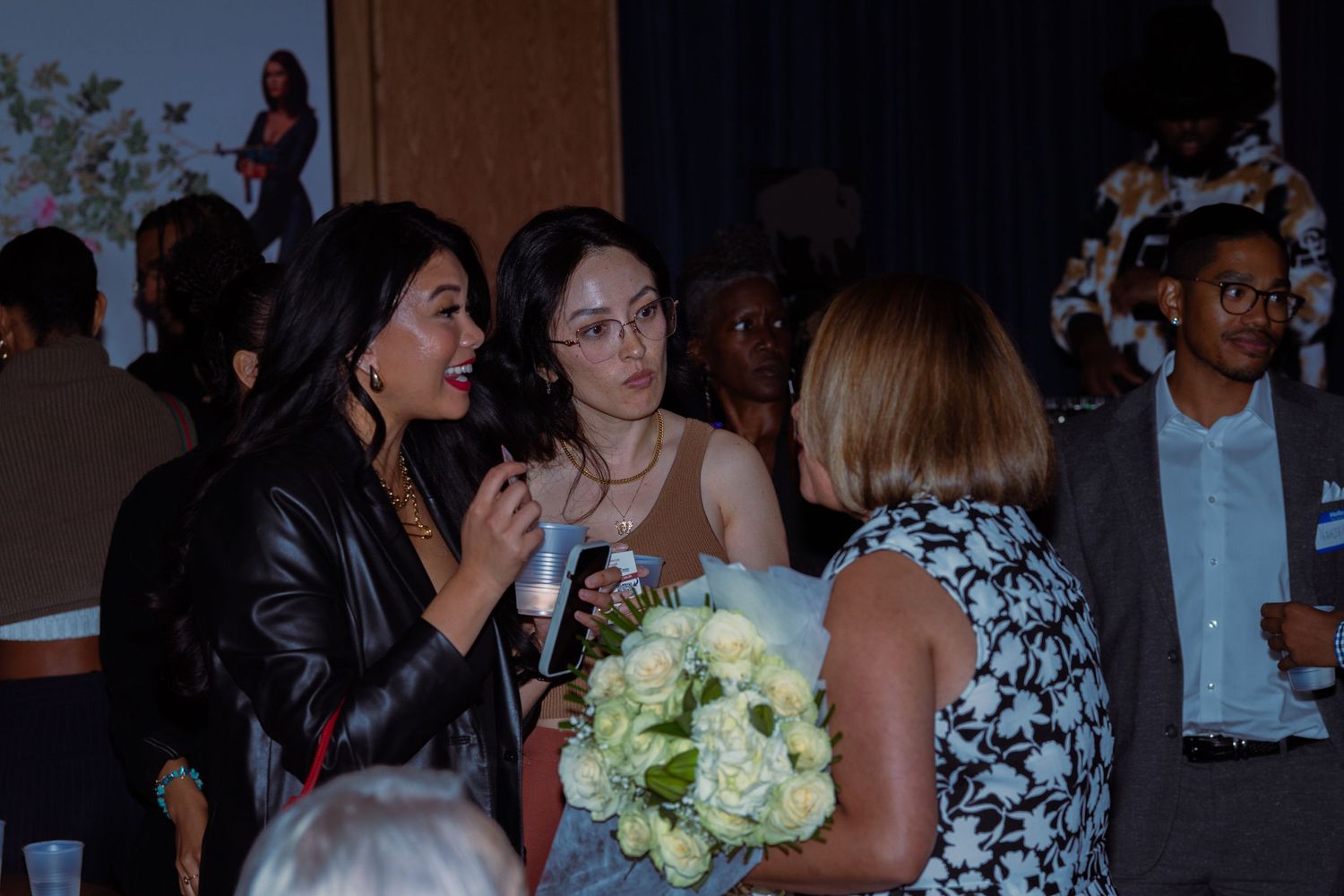 Women conversing at an indoor event, one holding white flowers, dressed elegantly in a warmly lit venue.