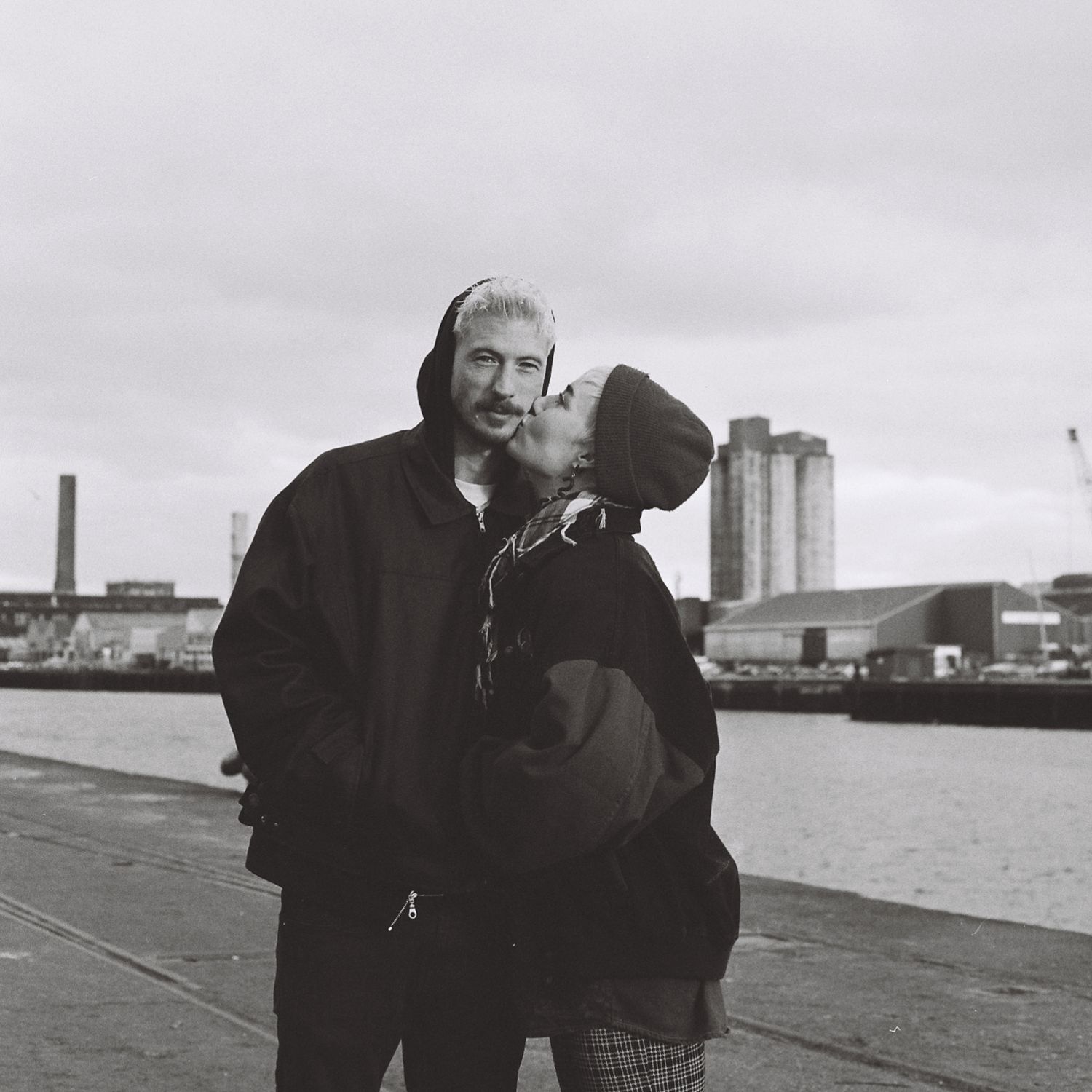 A romantic black and white photo of a couple embracing by the waterfront with industrial buildings in the background.