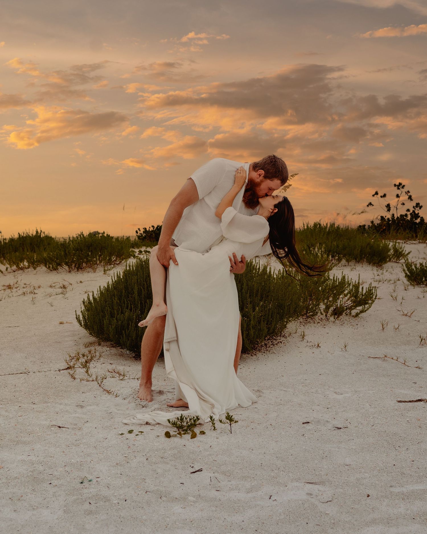 A romantic moment captured at sunset on a sandy beach with sea grass and glowing orange sky in the background.