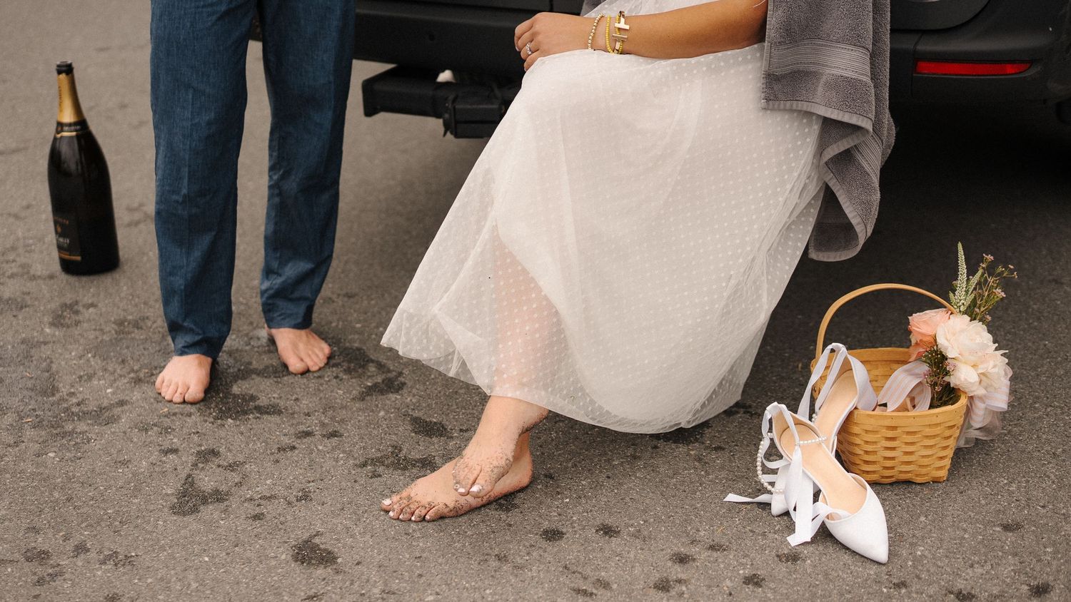 Bare feet stand beside a white lace dress, white shoes and a picnic basket with wine bottle on ground.