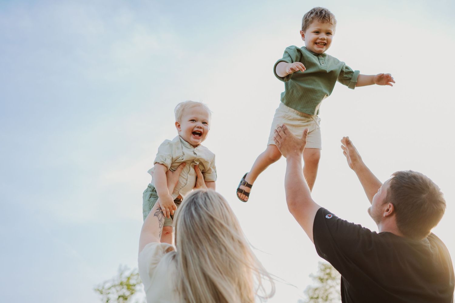 Joyful parents lifting and tossing two small children into the air against a bright blue sky.