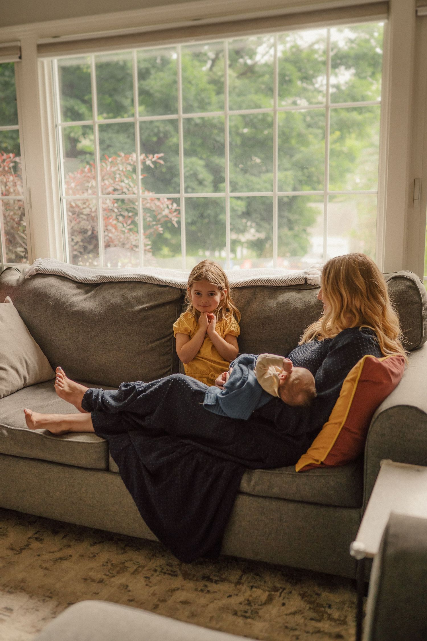 Family members relax together on grey sectional couch by bright windows.