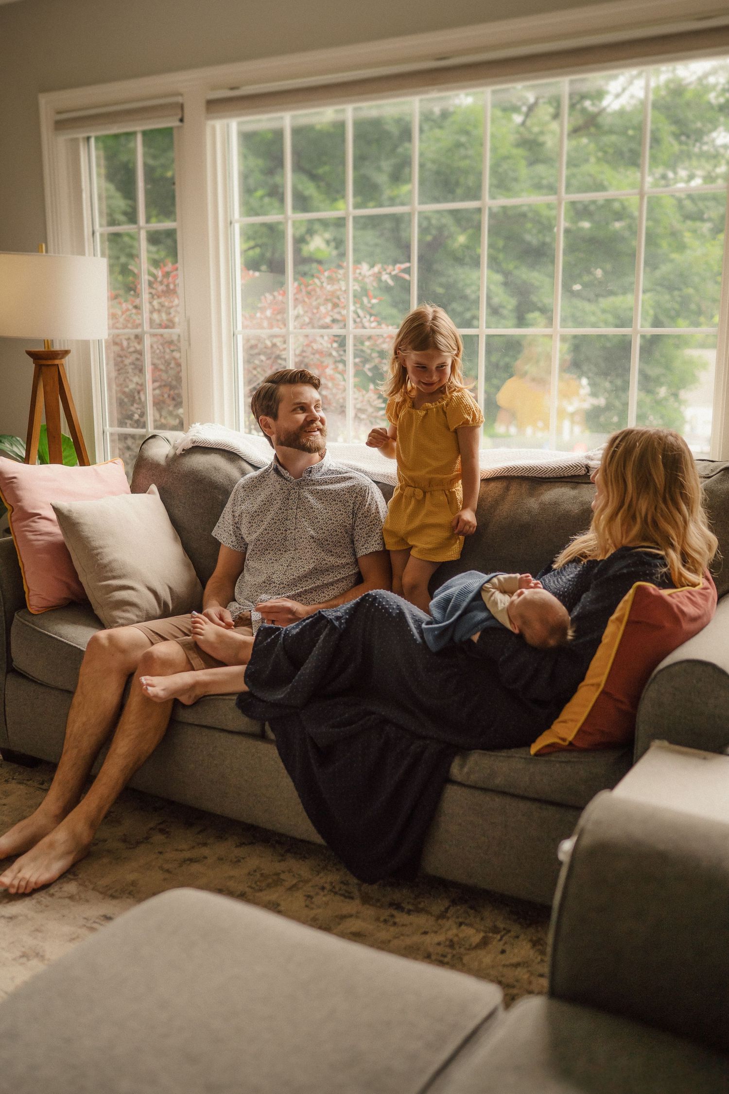 Multiple people relax together on a gray couch in front of bright windows in a living room.