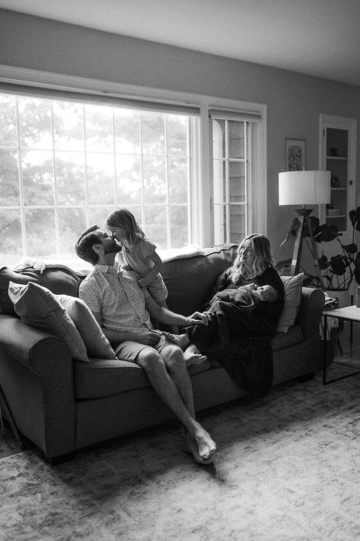 A black and white photo showing a cozy living room scene with people relaxing together on a sofa near a bright window.