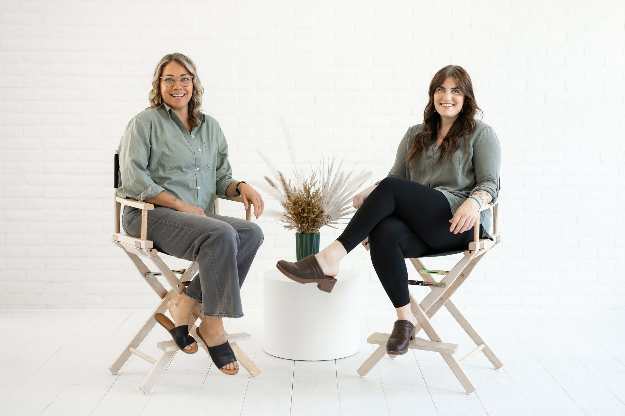 Two people sitting casually in wooden outdoor chairs against a white studio background.