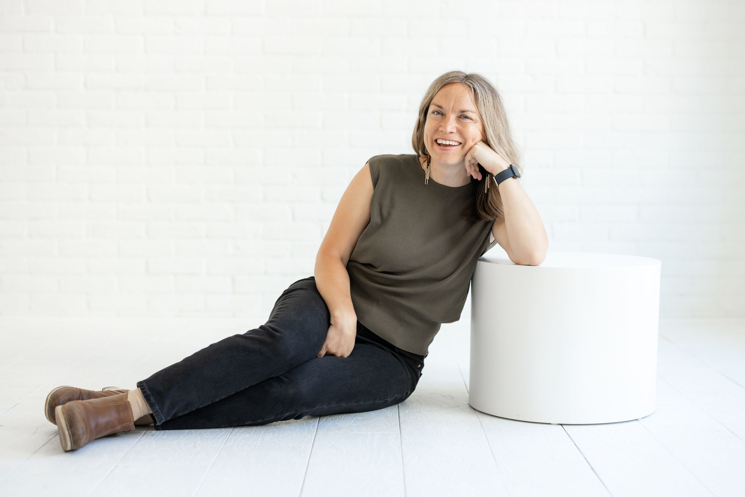 Someone in a sleeveless olive top and dark pants sits casually next to a white cylinder against a light background.