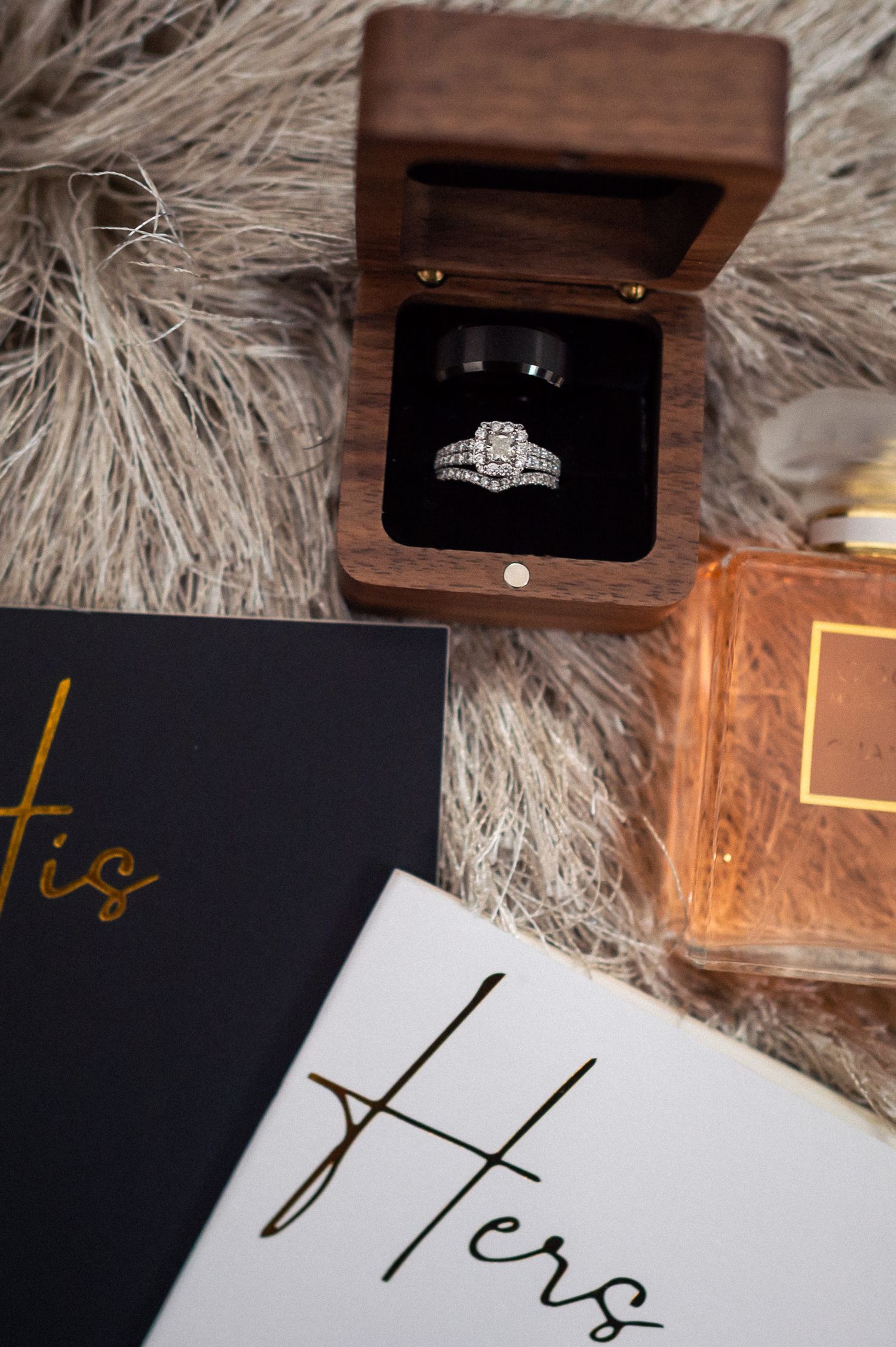 Diamond engagement ring in wooden box displayed with perfume bottle and 'His and Hers' wedding stationery on gray fur background.