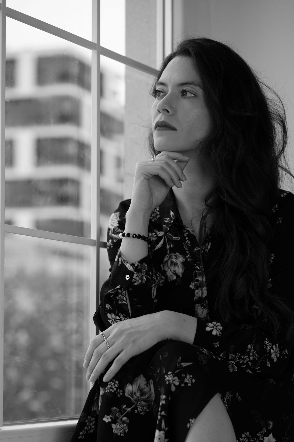 A woman in a black floral dress poses thoughtfully against a bright window in a moody black and white portrait series.