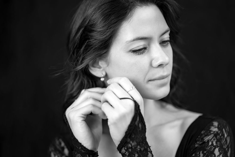 A black and white portrait of a person wearing a lace top, touching their ear with their eyes softly closed.