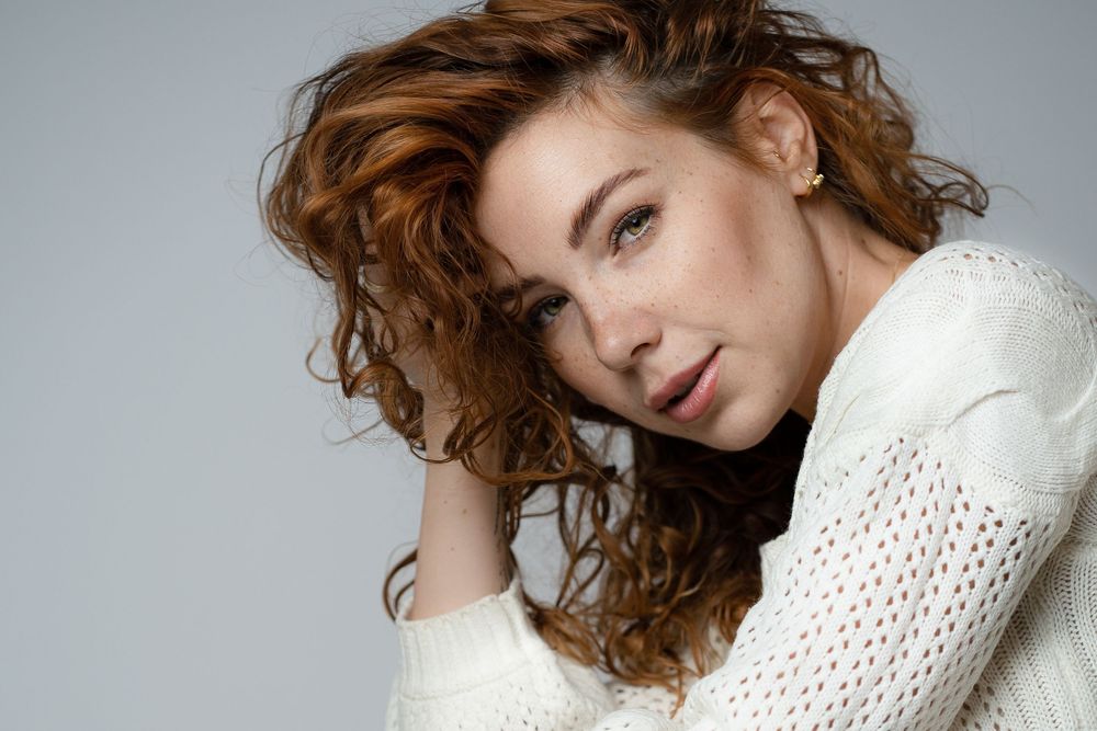 Fashion portrait featuring curly red hair and a white knit sweater against a light gray studio background.