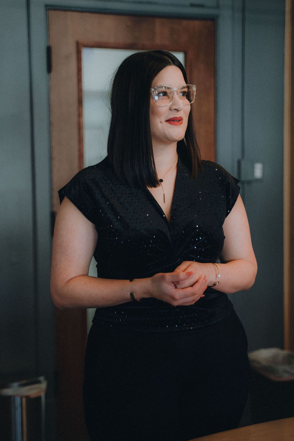 A professional wearing a black dress and glasses stands thoughtfully in front of a wooden door.