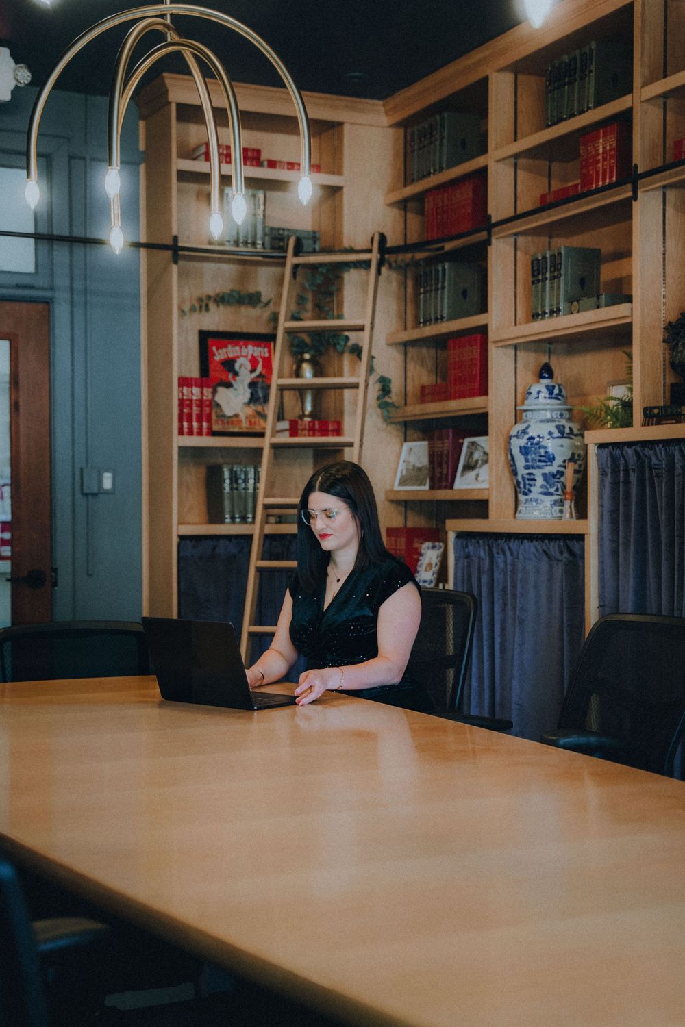 A person sits at a long wooden table in a library or office interior with bookshelves along the walls.