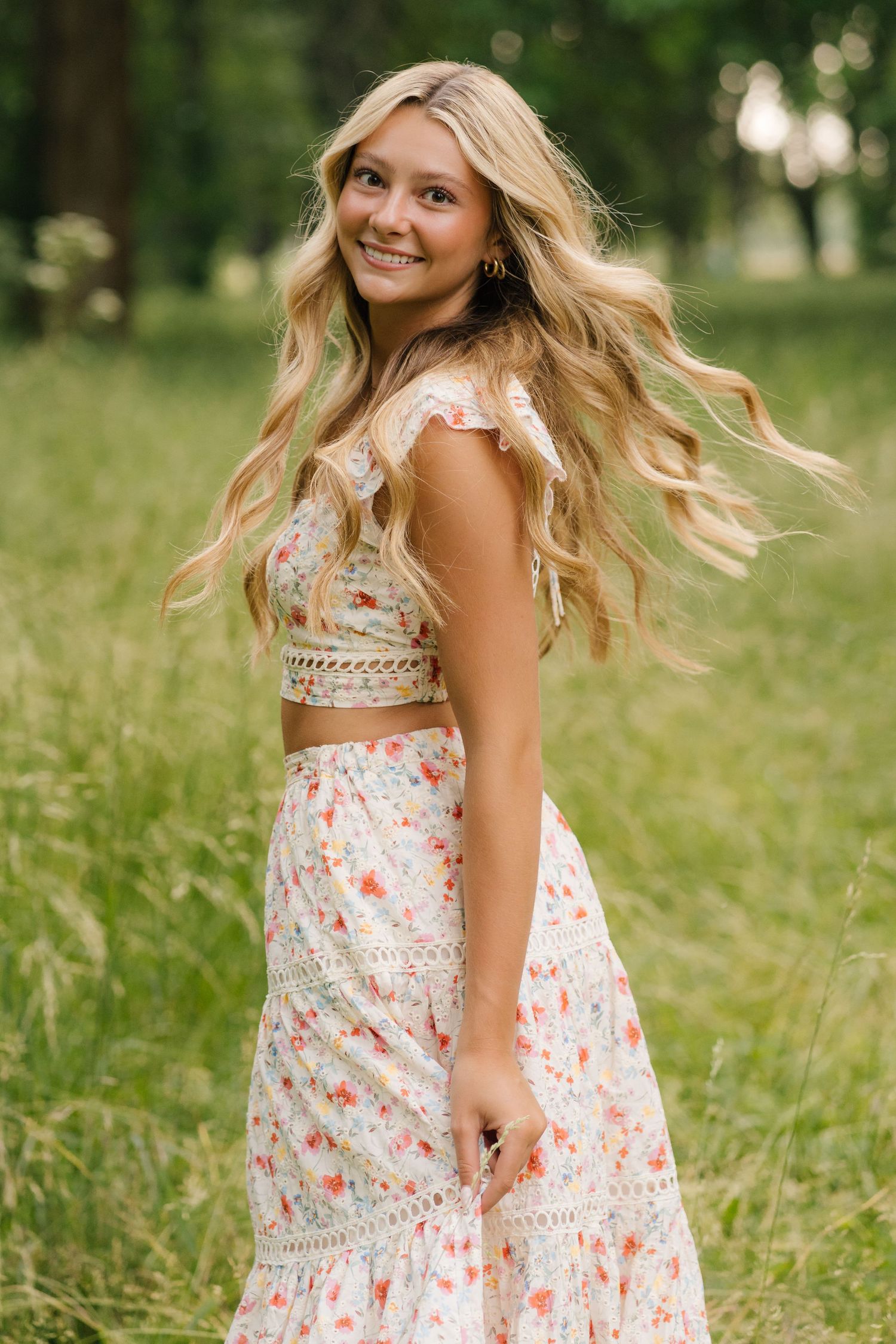 A young woman in a floral two-piece dress smiles while twirling in a sunlit meadow with flowing blonde hair.