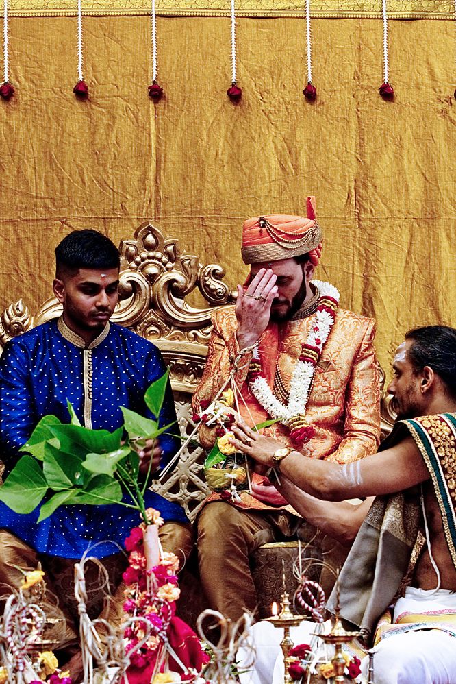 Traditional Indian wedding ceremony takes place against golden wall with ornate decorative throne and floral arrangements.