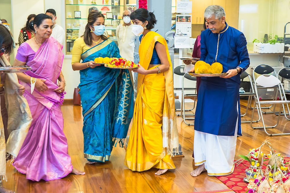 People in colorful traditional Indian saris and kurta carry bright yellow plates during a cultural celebration indoors.